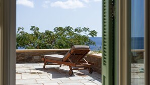Una silla de lounge de madera con ruedas en un patio de piedra con vistas al mar, con palmeras y un cielo despejado al fondo.
