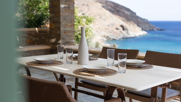 Mesa lista para comer con platos, vasos y un florero, con vista a un paisaje costero con un acantilado rocoso y el mar.