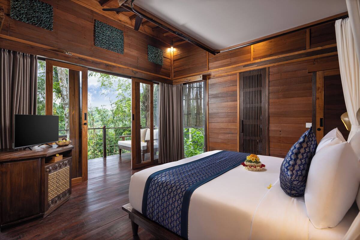 Ein Schlafzimmer mit einem großen Bett, Holzwänden, einem Fernseher auf einer Holzkommode und einem Balkon mit Blick auf Grünflächen.