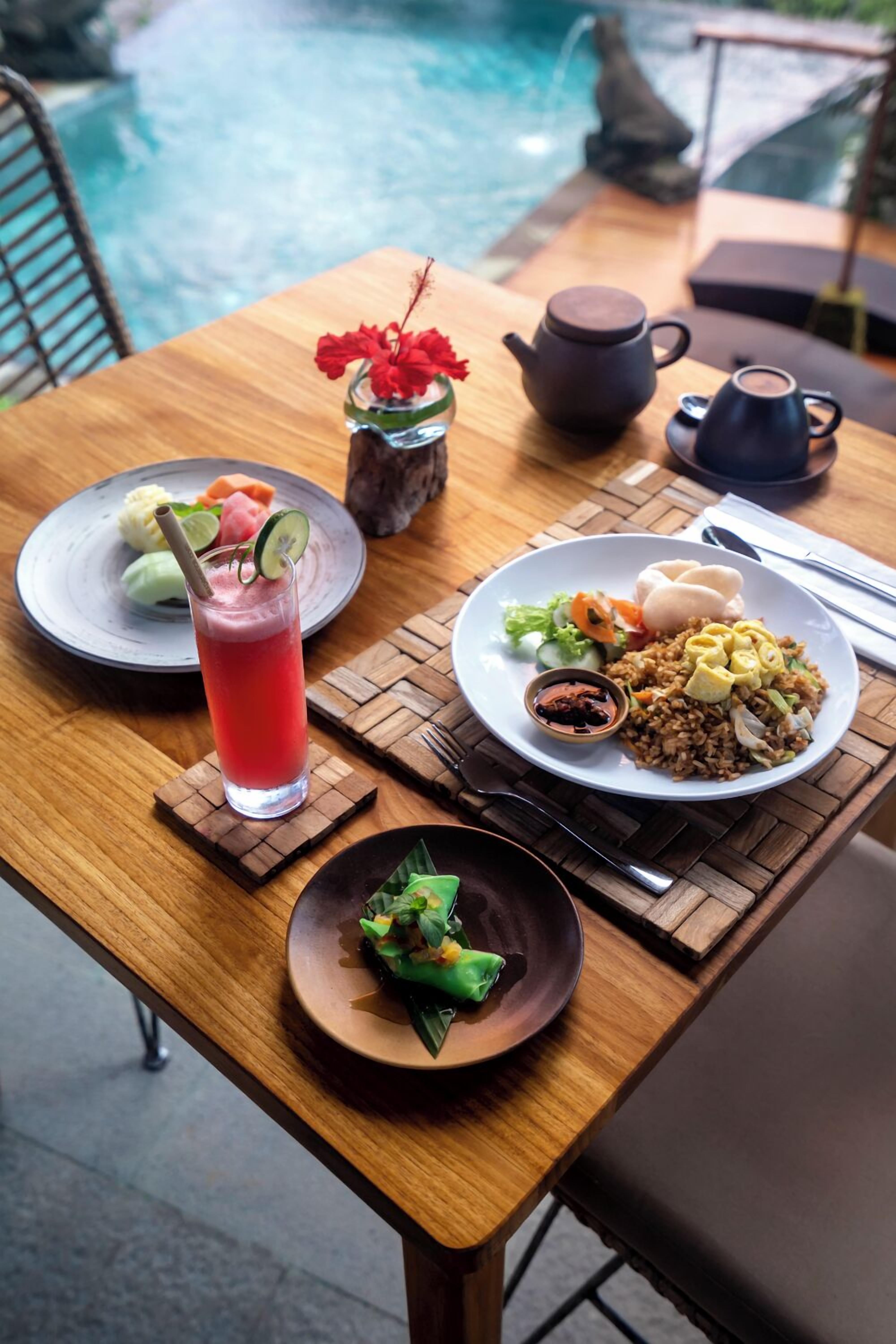 A dining table with a variety of dishes, a teapot, and a glass of red beverage, set against a backdrop of a pool.