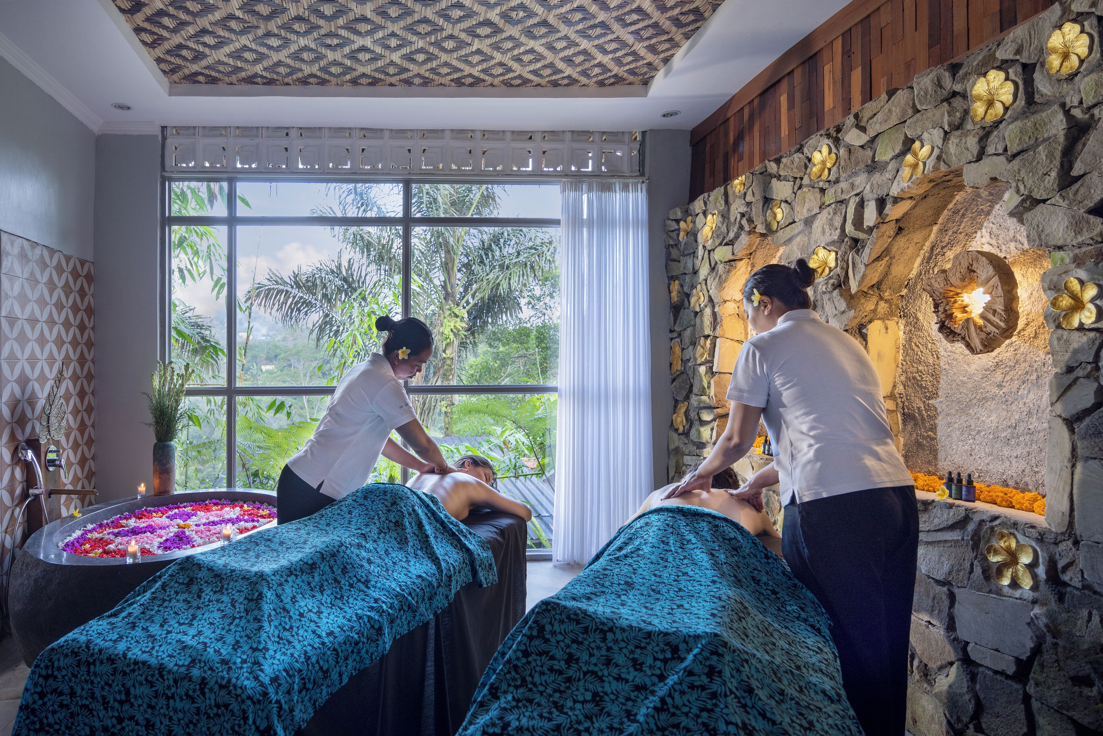 A spa room with two massage tables, a stone wall with floral decorations, and a large window with a view of greenery.