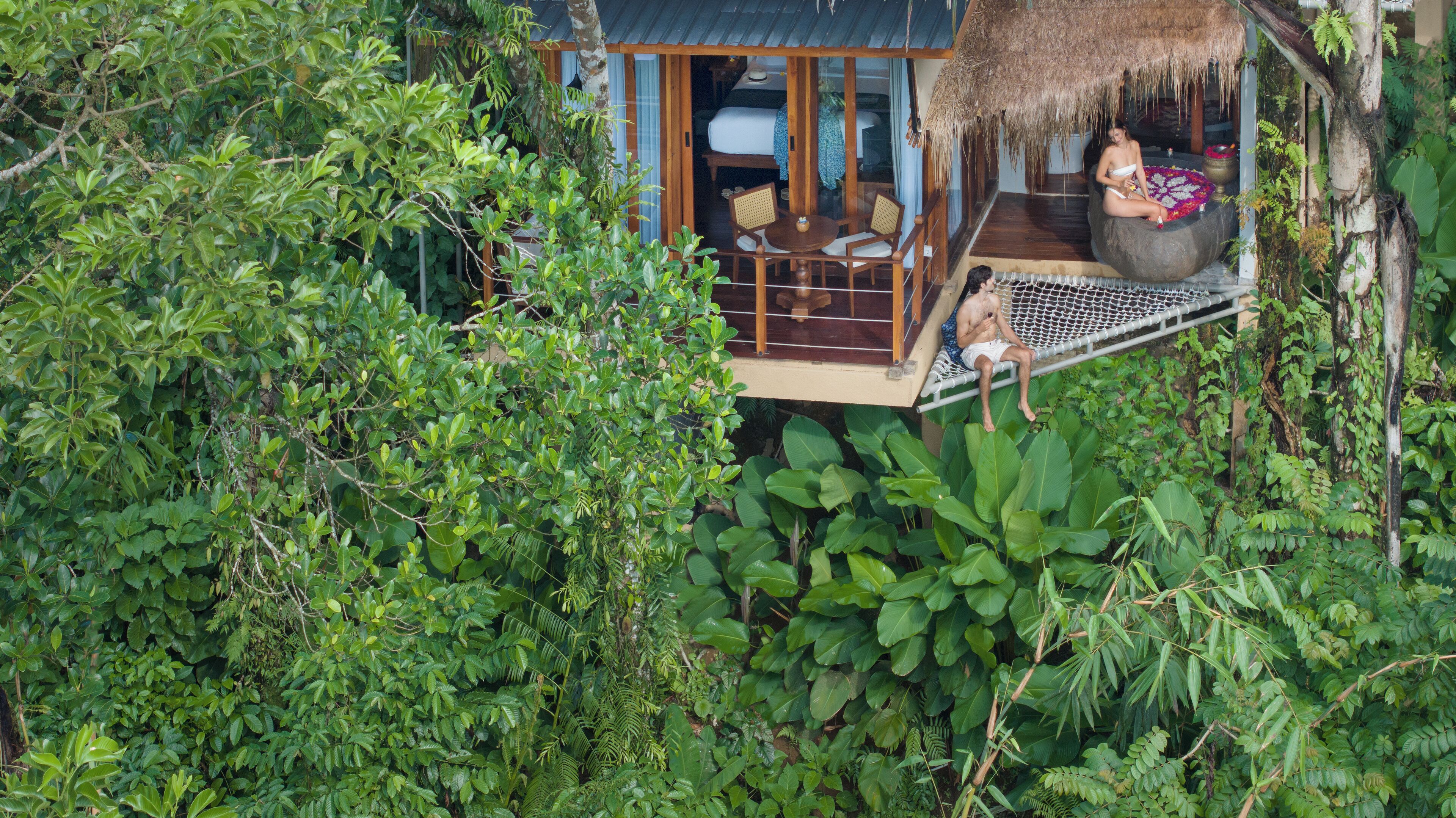 Une cabane perchée dans un arbre, avec un toit de chaume, une terrasse en bois et un petit espace salon comprenant une table et des chaises.