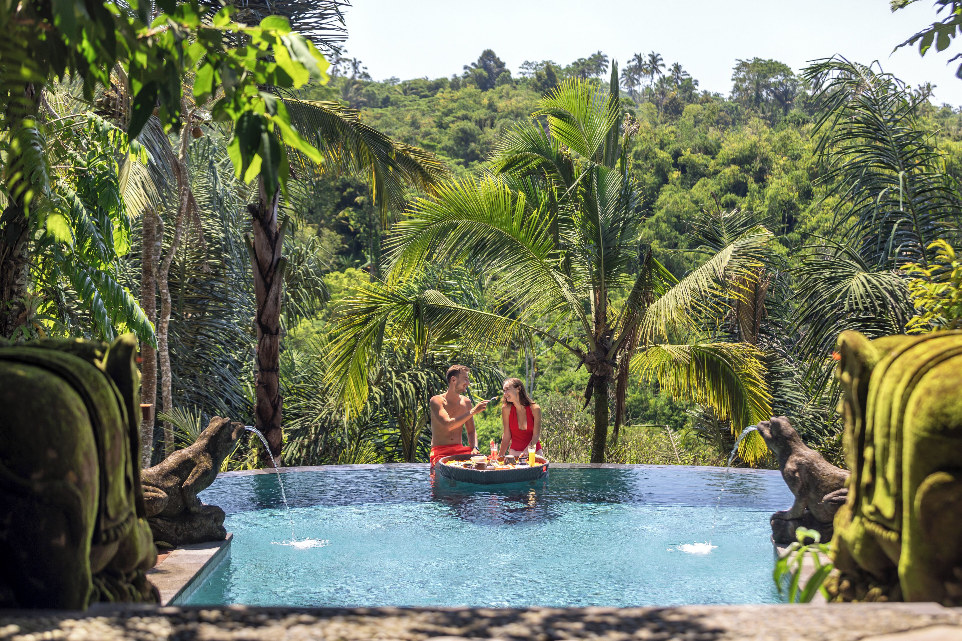 A pool with two people on a floating platform, surrounded by lush greenery and tropical plants.