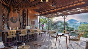 A wooden deck with tables and chairs, a bar area, and a view of a mountain. - Sacred Valley by GenuineHost (Sebatu)