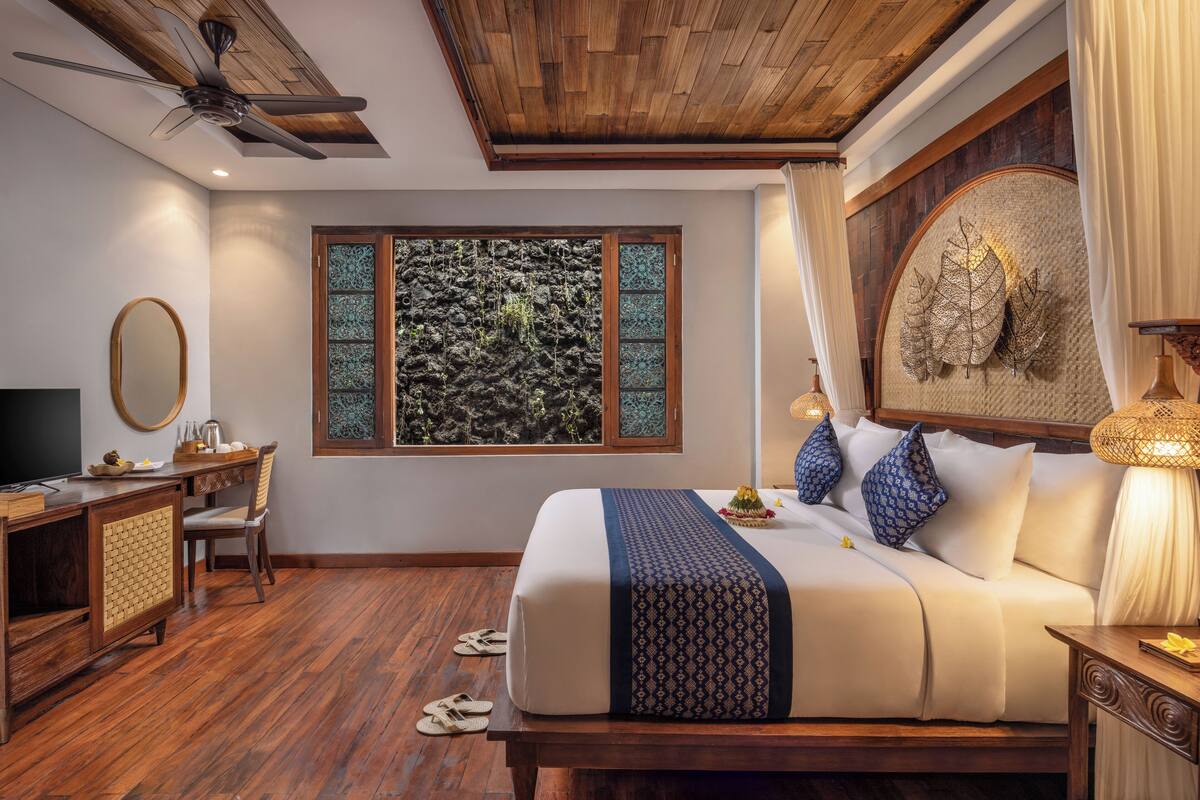 Ein Schlafzimmer mit Holzbalkendecke, einem Bett mit gemustertem Überwurf, einem Schreibtisch mit Spiegel und Blick auf eine felsige Wand durch ein Fenster.