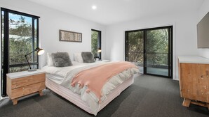 A modern bedroom with a large bed, a wooden nightstand, a desk, and a wardrobe. There are large windows and a view of trees outside. - Snowbound 2 - Family Ski Chalet, 5 minute walk to the slopes (Thredbo)