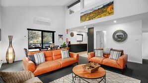 A modern living room with two orange sofas, a coffee table, and a dining area with a large window. - Snowbound 2 - Family Ski Chalet, 5 minute walk to the slopes (Thredbo)