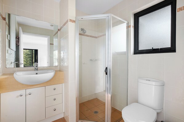 A bathroom with a white sink, mirror, and toilet, a glass shower enclosure, and a window with a black frame.