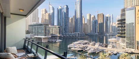A balcony with wicker chairs and a table, overlooking a marina with boats and a city skyline with modern high-rise buildings.