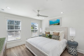 A bedroom with a bed, a ceiling fan, two windows with blinds, a nightstand with a lamp, and a painting on the wall.