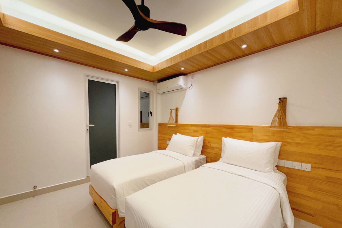 A room with two single beds, wooden headboards, a ceiling fan, and a small window.
