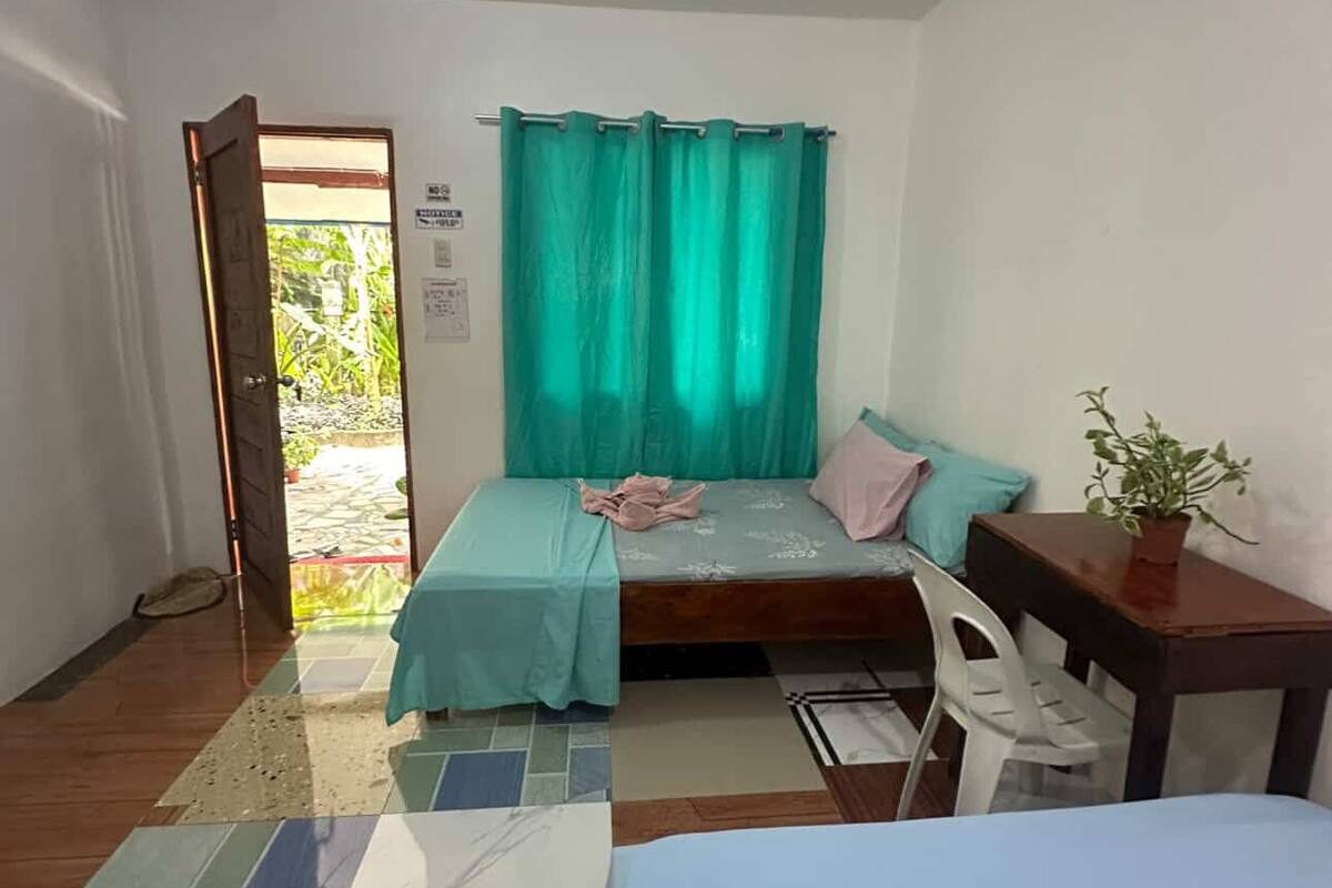 A small, well-lit room with a bed, a table, and a chair. There is a window with curtains, a door leading outside, and a tiled floor.