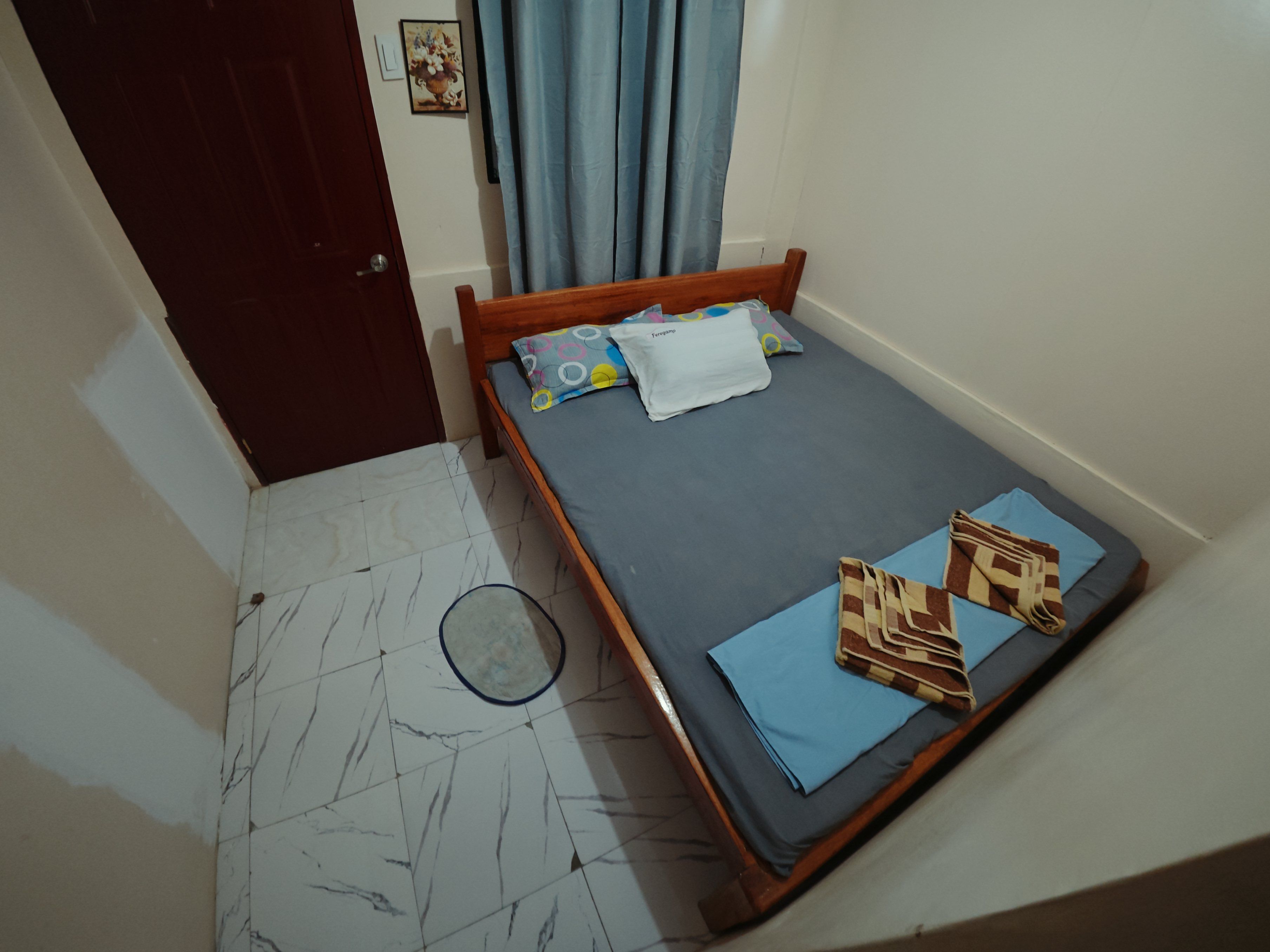 A small, single-bed room with a wooden frame, a grey mattress, and a blue curtain.