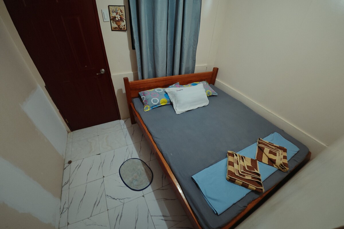 Une petite chambre avec un lit simple, un cadre en bois, un matelas gris et un rideau bleu.