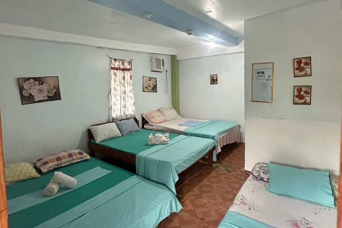 A room with three beds, each with a green bedspread, a window with floral curtains, and a wall-mounted air conditioning unit.