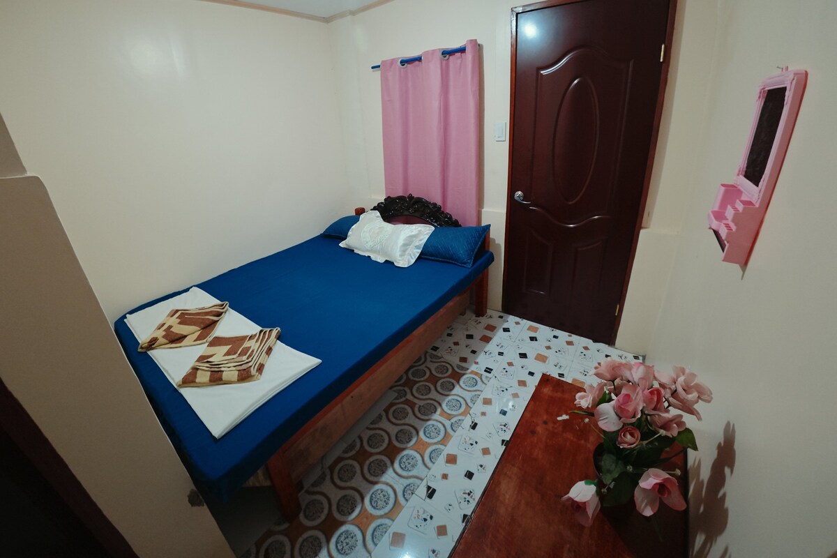 Une chambre avec un lit simple, une porte en bois, une petite table sur laquelle est posé un vase de fleurs et un sol à motifs.