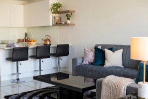 A modern living room with a grey sofa, black chairs, a coffee table, and a kitchen area with white cabinets and a kettle.