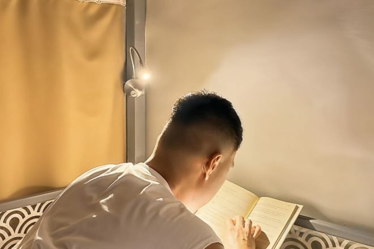 A person reading a book in a room with a bed and a bedside lamp.