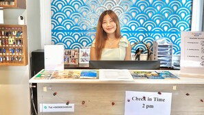 A reception desk with a blue patterned backdrop, a sign indicating check-in time at 2 pm, and a display of brochures and a telephone number. - Friends Hostel - Phuket Airport (Sa Khu)