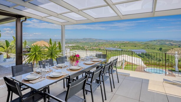 A dining area with a table set for four, overlooking a scenic landscape with hills and a pool. - Beautiful villa with pool, SPA and panoramic sea views, just 10 minutes from the beaches of Pineto (Atri)