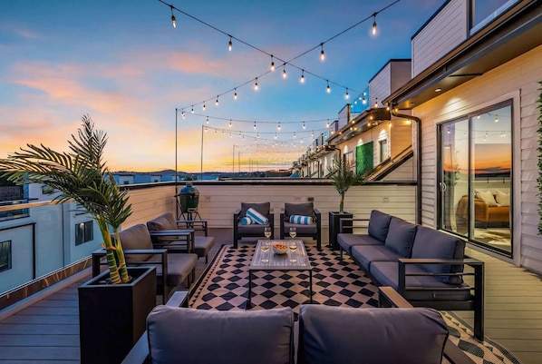 A rooftop terrace with outdoor seating, string lights, and a view of the sunset.