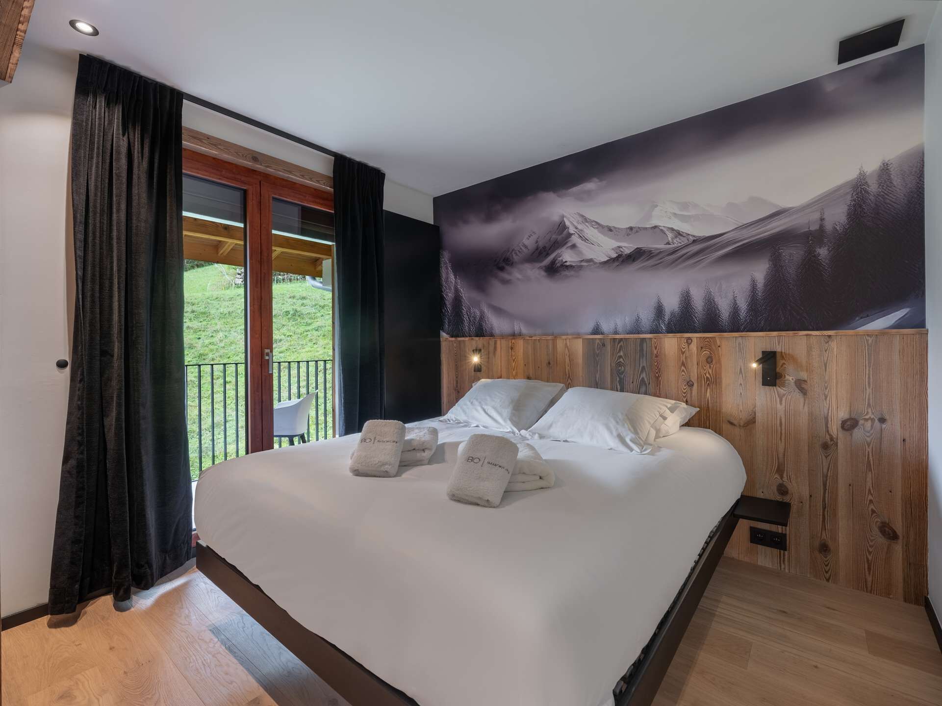 Un dormitorio con una cama grande, paneles de madera, un mural con paisaje de montaña y un balcón con una silla.