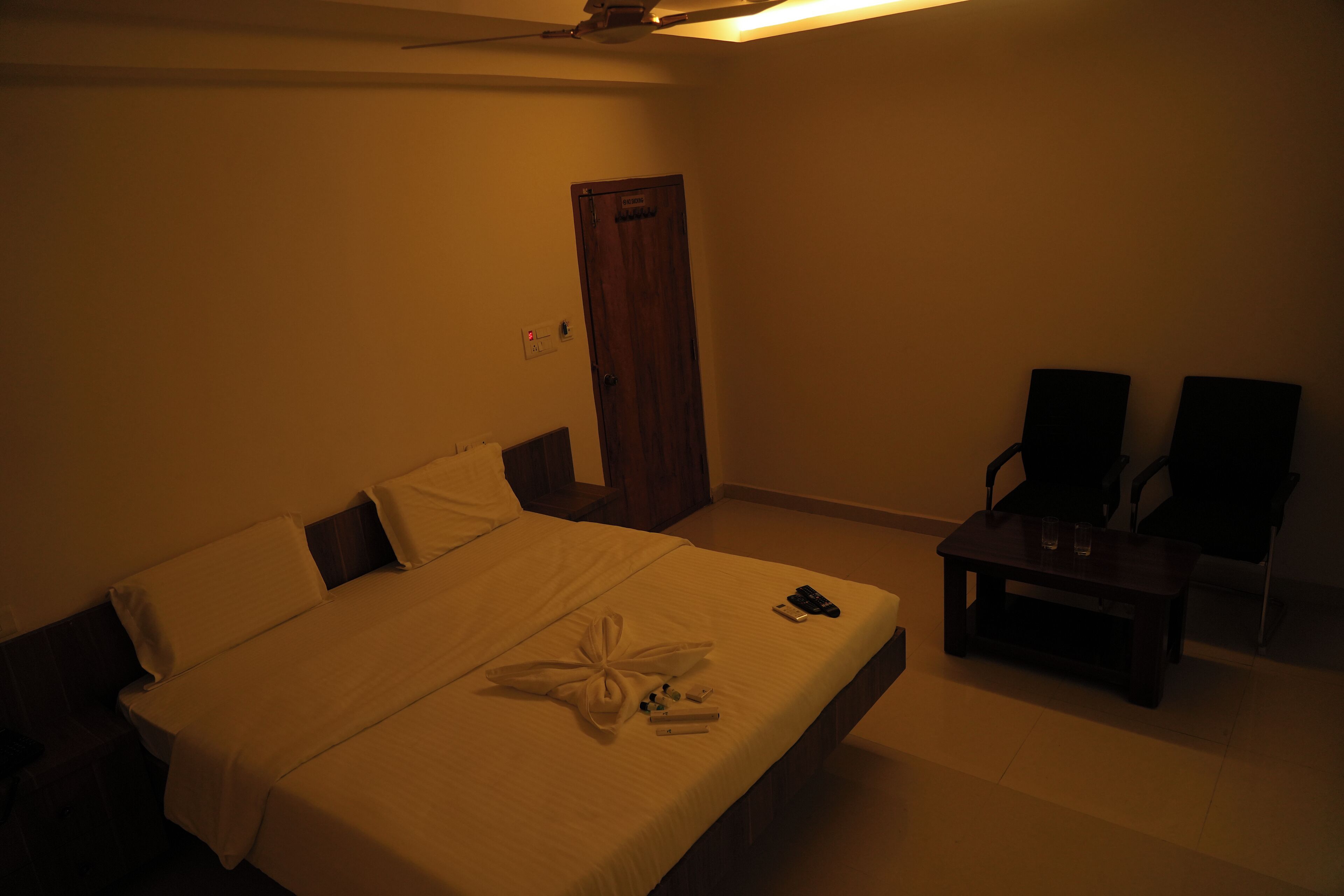 A dimly lit hotel room with a bed, bedside table, and two chairs.