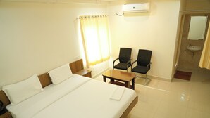 Une chambre d’hôtel avec un grand lit, deux chaises, une petite table, une table de chevet, une fenêtre avec des rideaux, une salle de bain avec un lavabo et un miroir, et un appareil de climatisation.