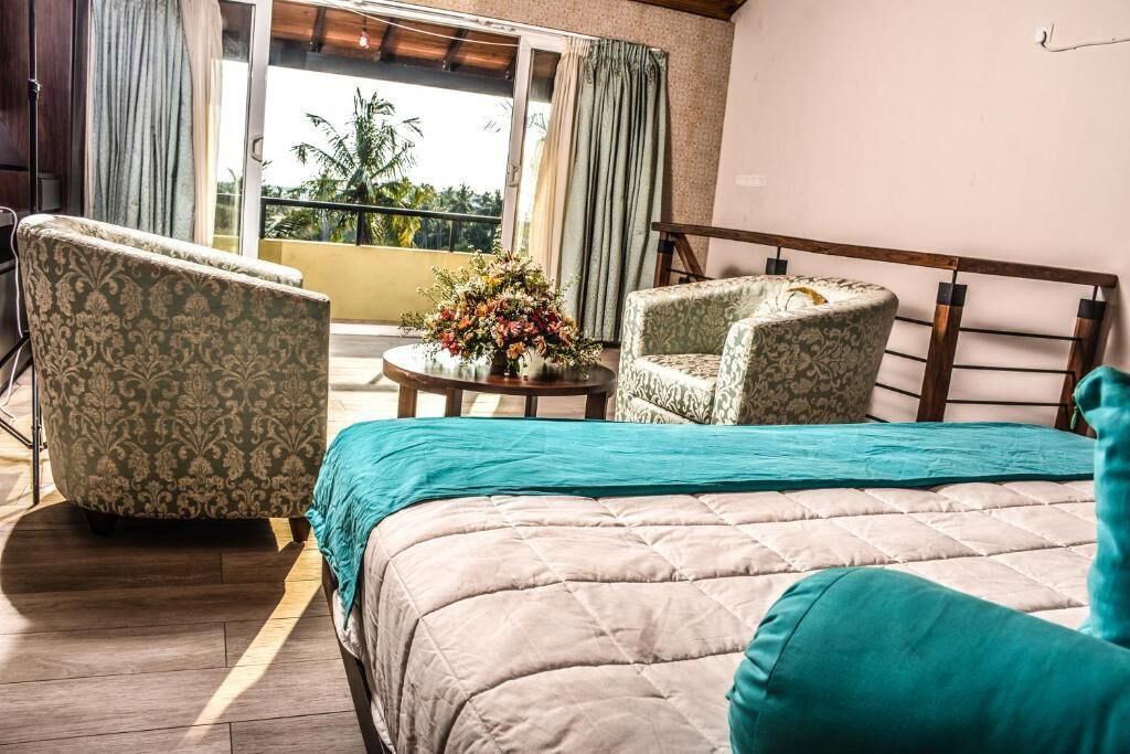 A bedroom with a bed, two patterned armchairs, a small table with a flower arrangement, and a balcony with curtains.
