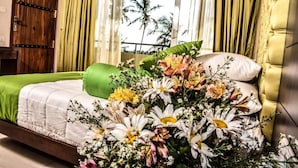 A bedroom with a bed, green and white bedding, a floral arrangement, and a view of palm trees through a window.