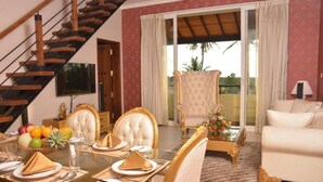 A dining area with a glass table set for four, surrounded by ornate chairs. A staircase is visible to the left, and a balcony with a view of palm trees is in the background.
