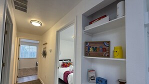 Ein Flur, der zu einem Schlafzimmer führt, mit einem Bett, einem Regal mit Büchern und einer gelben Tasse, sowie einer Tür mit Fenster.