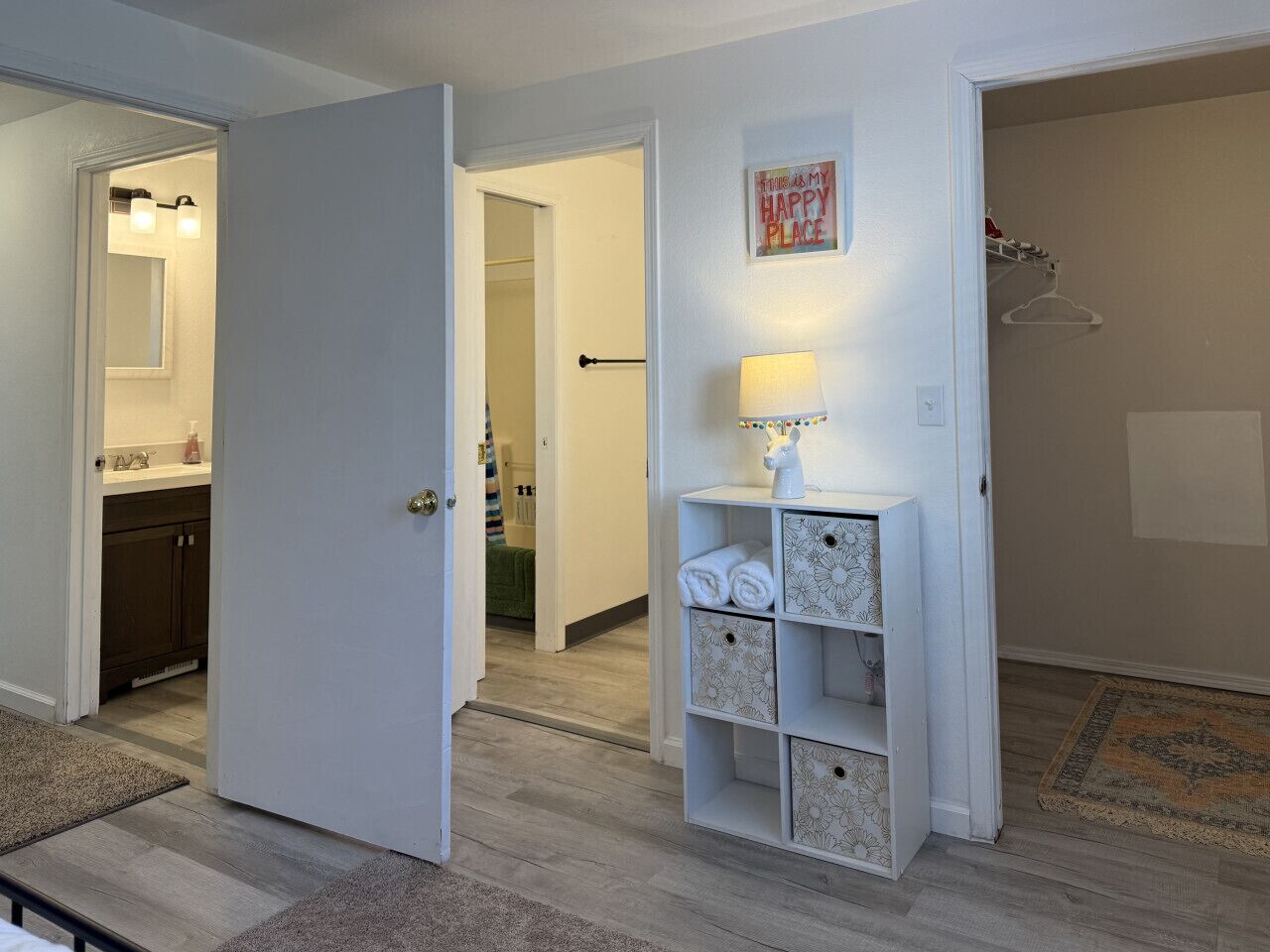 Un pasillo con una puerta blanca que da a un baño, un estante pequeño con una lámpara, y una alfombra en el suelo.