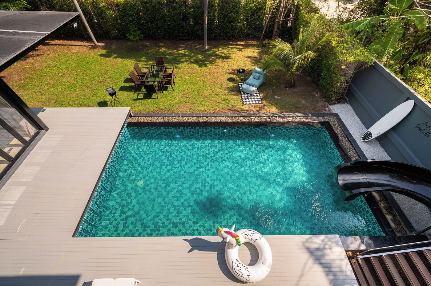 Piscina transparente com um deque de madeira, cadeiras de descanso e uma prancha de surf.