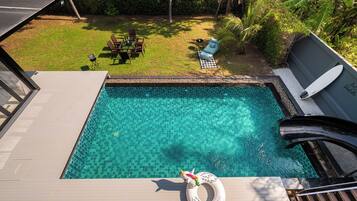 Piscina transparente com um deque de madeira, cadeiras de descanso e uma prancha de surf.