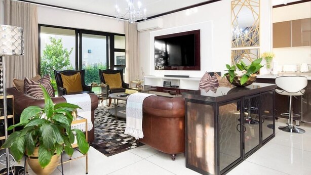 A modern living room with a sofa, coffee table, and TV. There is a large window, a chandelier, and a plant. - Craigavon House by Tanaka Kingdom  (Fourways)