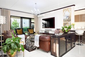 A modern living room with a sofa, coffee table, and TV. There is a large window, a chandelier, and a plant.