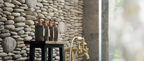 A bathroom with a stone wall, a wooden shelf with toiletries, a gold faucet, and a white bathtub.
