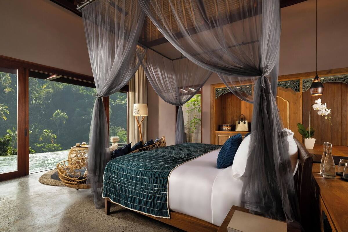 Een ruime slaapkamer met een groot bed, een houten nachtkastje, een rieten stoel en uitzicht op een tropisch landschap.