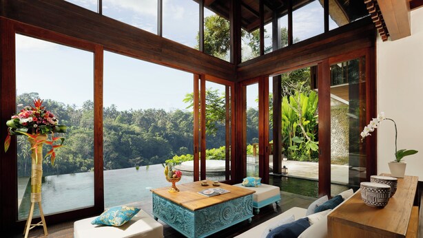 A spacious room with a large window overlooking a lush landscape. There is a wooden coffee table with a vase of flowers, a sofa with cushions, and a side table with a potted plant.
