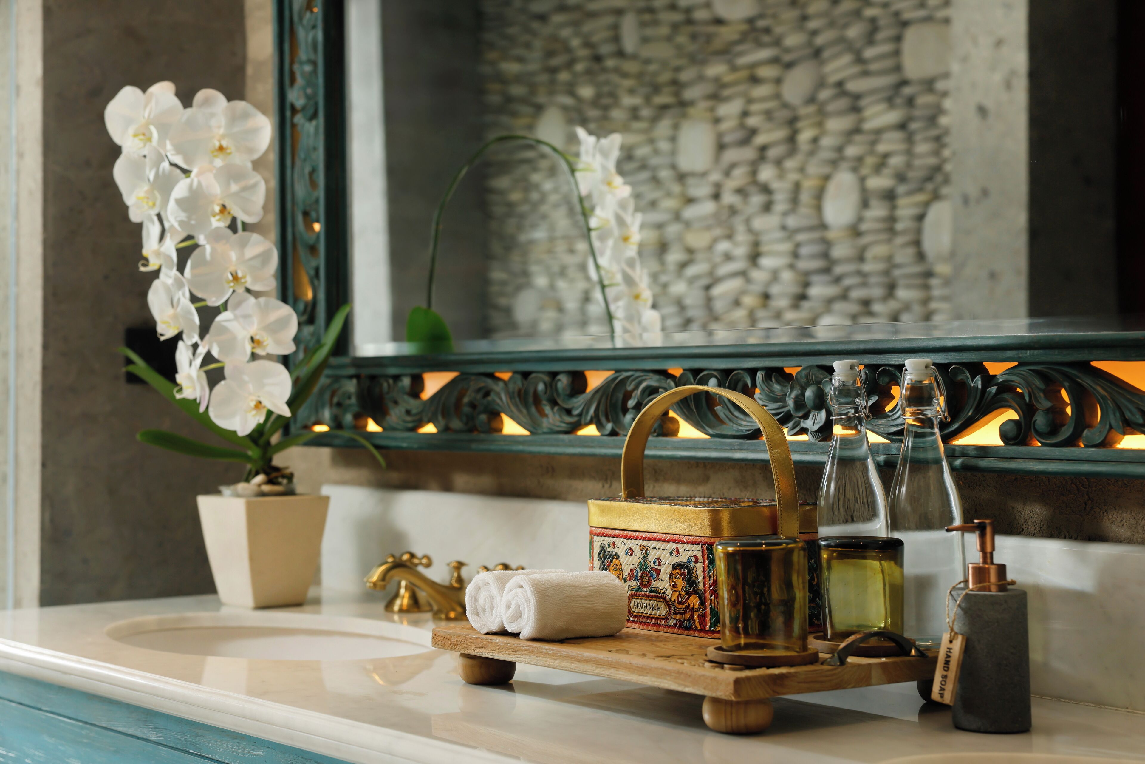A bathroom vanity with a mirror, a potted orchid, a towel, a decorative tray with toiletries, and a stone soap dispenser.