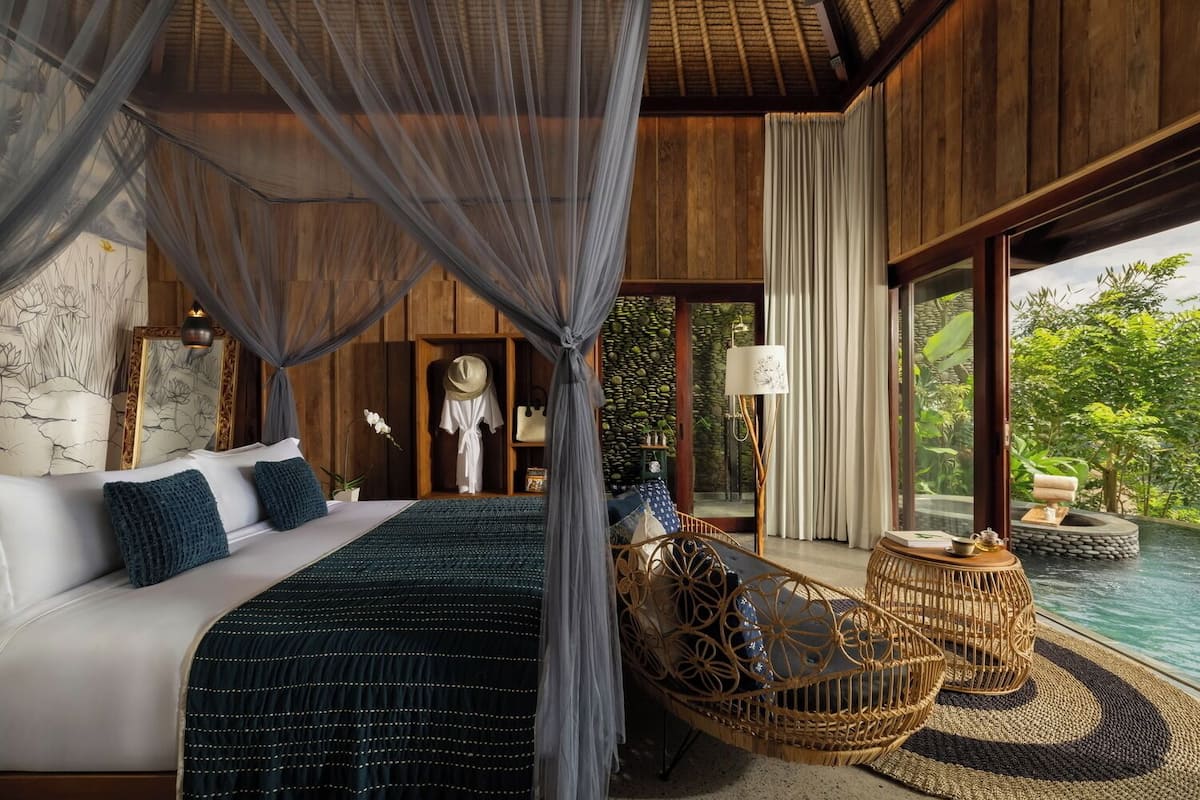 Een luxueuze slaapkamer met een hemelbed, een rieten stoel en uitzicht op een zwembad.