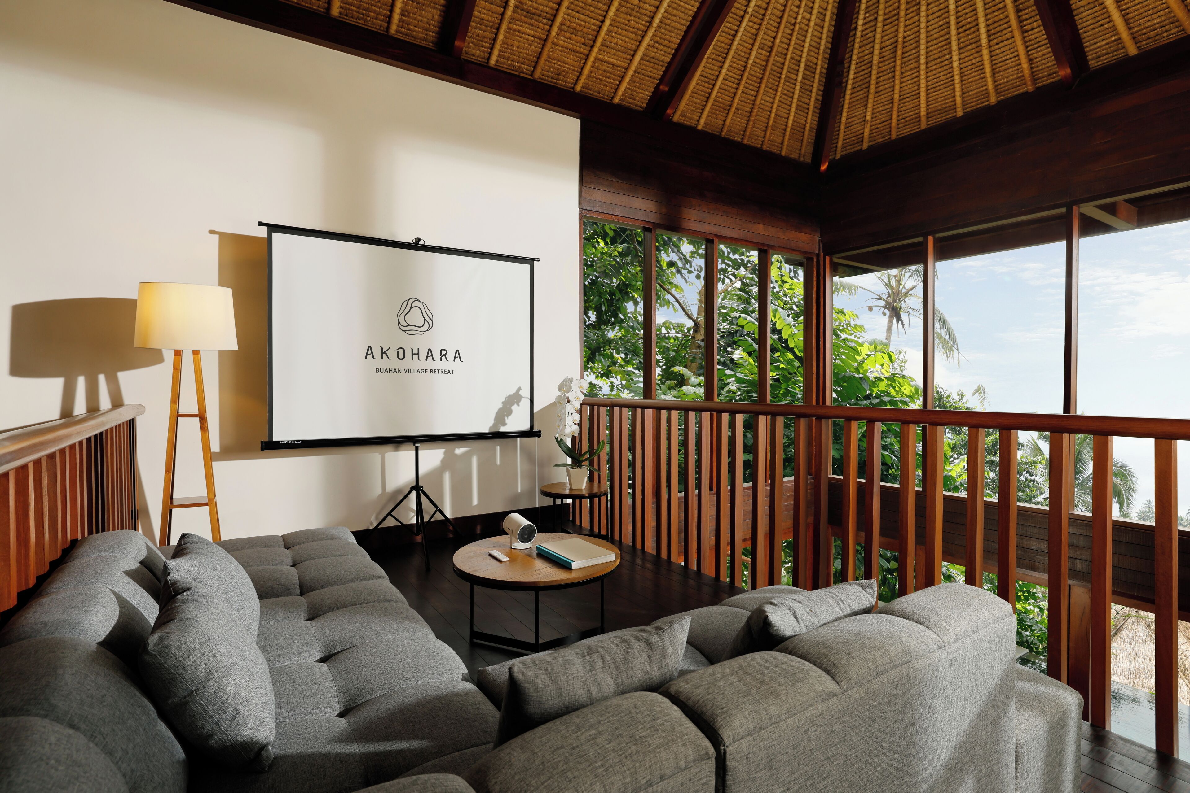 A modern living room with a large gray sectional sofa, a small round table with a lamp, and a projector screen displaying "AKOHARA BUAHA Village Retreat".