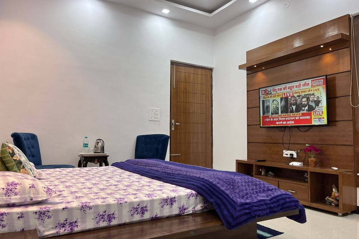 A bedroom with a bed, two chairs, a TV mounted on the wall, and a wooden wardrobe.