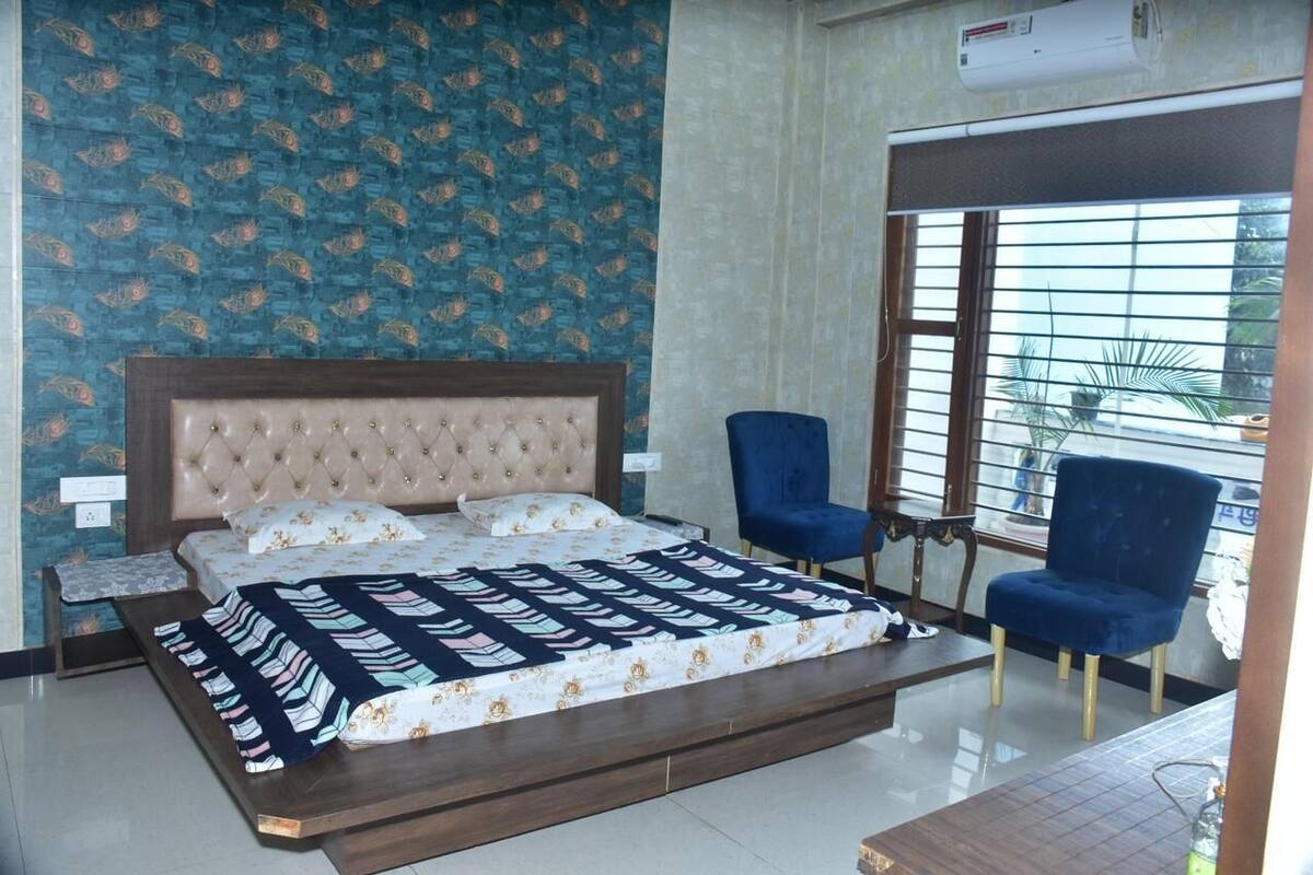 A bedroom with a bed, two blue chairs, a window with blinds, and a wall air conditioner.