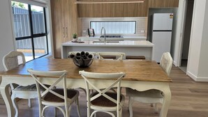 A modern kitchen with a wooden dining table, white chairs, and a central island with a sink and stove. - The Cosy Cottage  (Ararat)