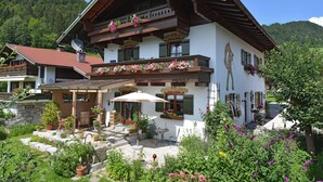 A traditional chalet with a balcony adorned with flowers, a patio with outdoor furniture, and a mountain backdrop.