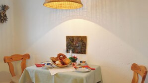 A dining table with a light green tablecloth, wooden chairs, a basket of pretzels, a small plant, and a painting on the wall.
