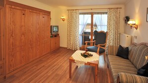 A cozy living room with wooden flooring, a sofa, a dining table with a lace tablecloth, a wooden cabinet, a television, and a window with curtains.