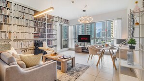A modern living room with a large bookshelf, a flat-screen TV, a dining area, and a view of high-rise buildings. - 2BR w Burj Khalifa View near Dubai Mall (Dubai)